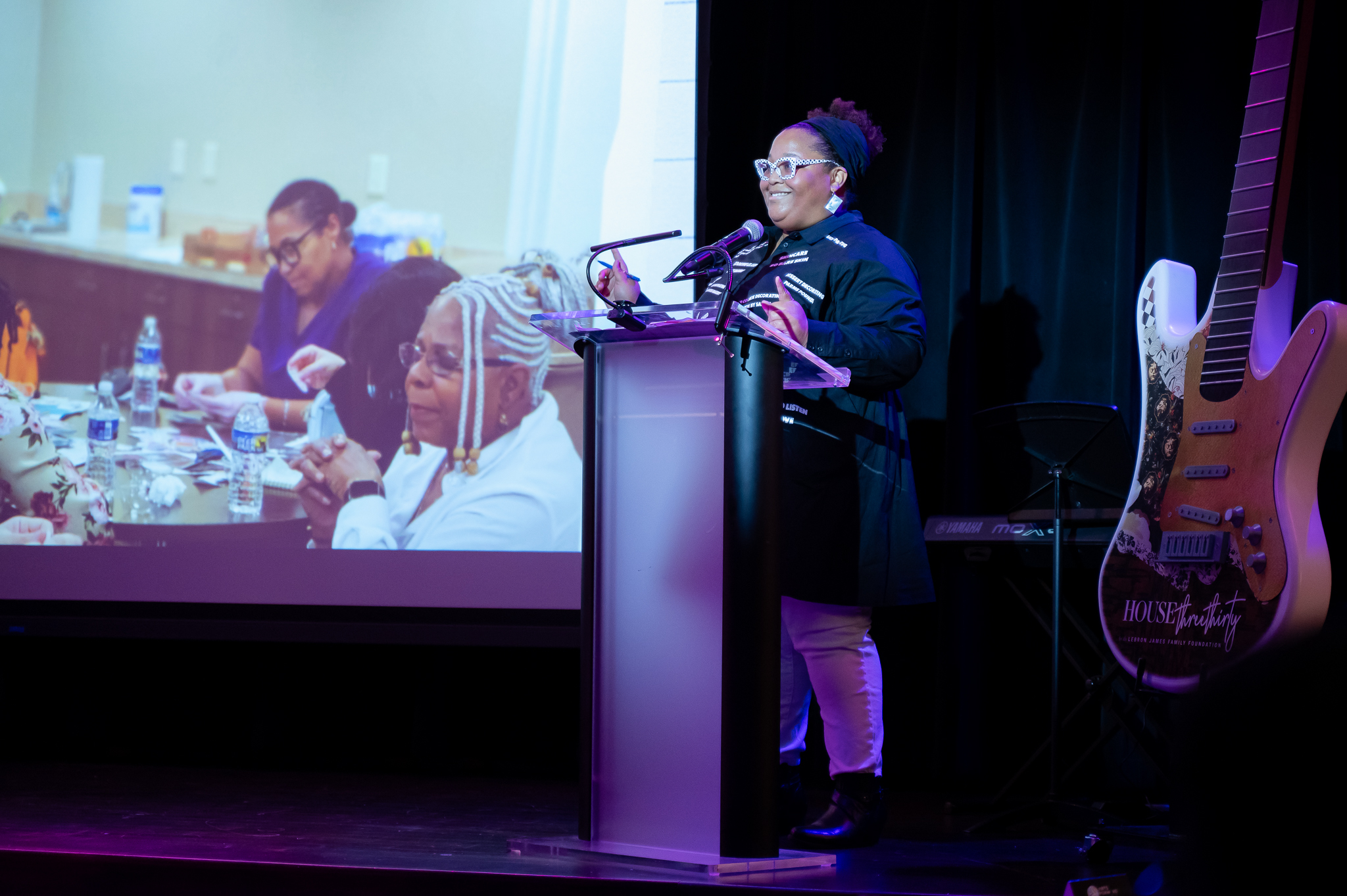A woman presenting on stage with a guitar behind her and presentation in the background.