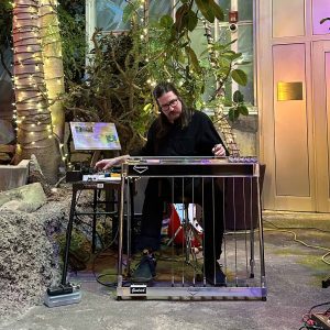 Photo of a man playing various electronic instruments in front of greenery