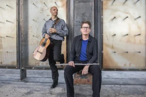 Two men in front of a wall, one holding a guitar and one holding a flute