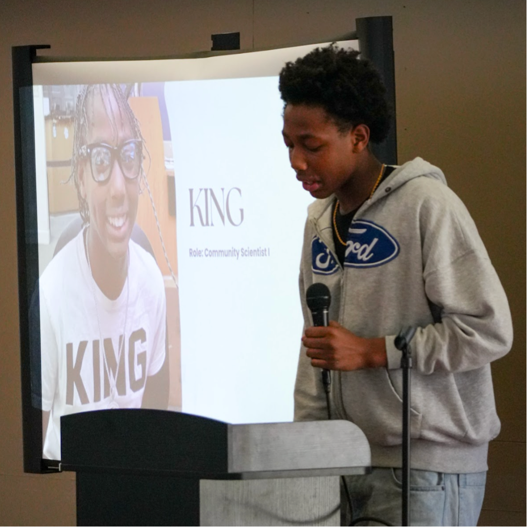 Blueprint student in a light grey Ford hoodie holding a microphone as he gives a speech.