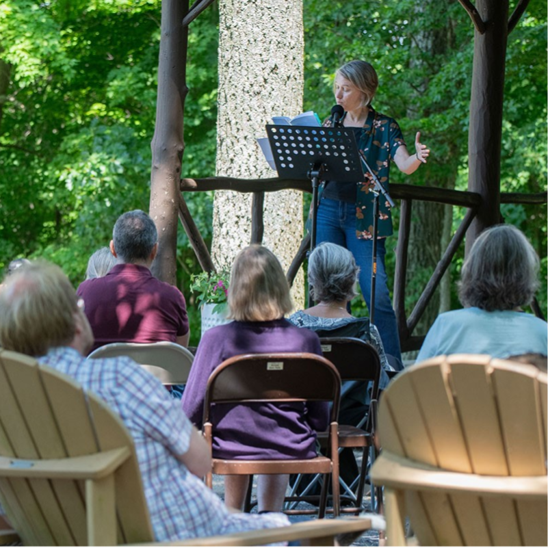 A white woman reading from a music stand in front of a forested area with audience member in adirondack chairs