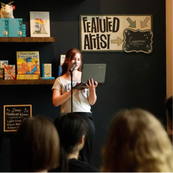 A white teenager reading from a laptop screen to an audience in a bookstore