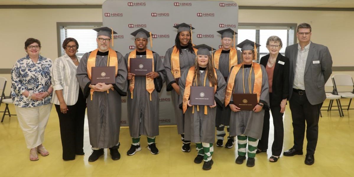 A group of adults of different ages and races pose in graduation gowns with their degrees.