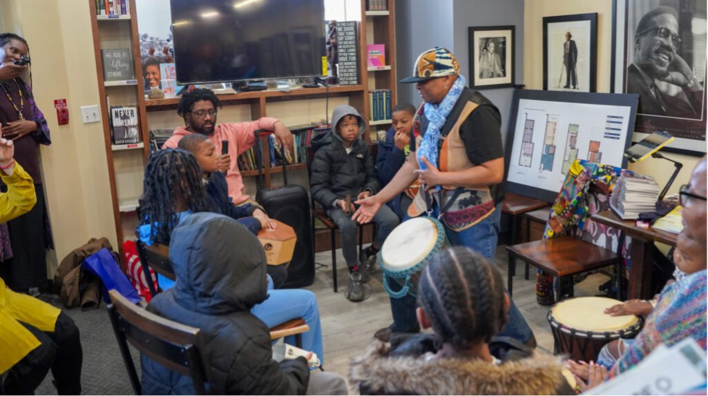 Black adults, and children gathered around a person with a drum in a living room setting.