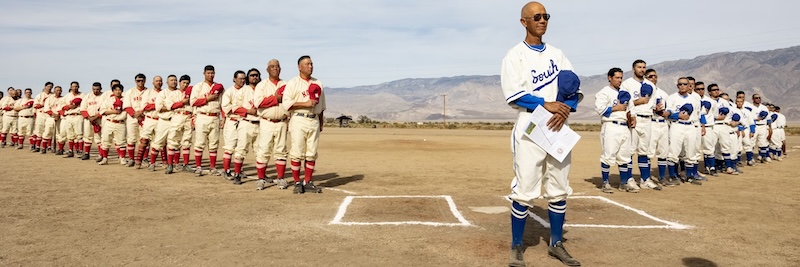A photo of Manzanar Baseball Project lead artist Dan Kwong standing in front of two baseball teams for a 2024 exhibition game at Manzanar. Many of the baseball players are descendants of internment camp detainees. They wear custom 1940s-style uniforms.