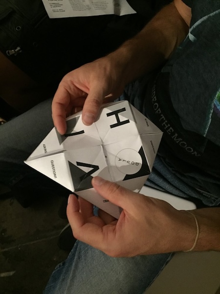 A photo of hands holding a folded paper fortune teller. The fortune teller is white paper printed with black text. The letters HIV are visible in the middle. 